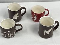 Four Starbucks mugs; three brown with animal and heart designs, one red with animal design shown from front on white cloth background.