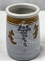 Front view of cylindrical pottery utensil holder vase with grape and leaf design and brown band near top.