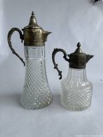 Two glass and metal carafes with diamond cut patterns and hinged metal lids standing side by side on a white background.