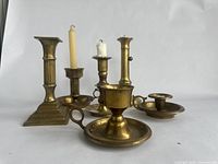 Photo showing all six brass or metal candlesticks arranged upright, highlighting their varying designs and wax remnants.