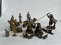 Overview of approximately 17 assorted cultural and mythological metal figurines arranged on a white surface. Includes standing figures, animals, cherub, and abstract shapes in metallic bronze and gold tones.