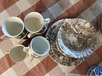 Photo showing 4 white cups with handles alongside stacked plates and bowls with brown Cotswold pattern on white ironstone.