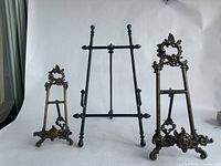 Three metal display stands standing together on a white background - two are ornate brass, one is black with simpler design.