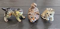 Photo showing three cat figurines on a dark surface, including a striped cat, a brown detailed cat, and a small light cat with stripes.