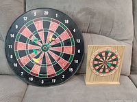 Photo showing both dart boards side by side on a couch, magnetic board on left with yellow and green magnetic darts, cork board on small wooden stand on right.