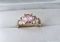 Front view of gold-tone ring showing pink square center stone and clear round side stones, placed in white cushioned ring holder.