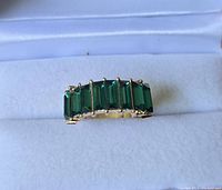 Close-up photo of the fashion ring showing seven rectangular green stones set in gold-toned band on white fabric.