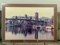 Framed cityscape print with city buildings, bridge, sailboats, and water reflection, housed in a metal frame with matting and glass protection.