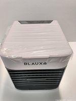 Photo of the Blaux Desktop AC Ultra unit wrapped in plastic, showing the white top with control panel and black front air vent grille.
