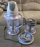 Photo shows Ninja blender motor base attached to a large plastic processing cup with handle, along with a smaller plastic processing cup, each with blade attachments. The blender is corded electric with a standard power cord.