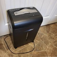 Black Insignia paper shredder with silver panel on top, positioned in front of white door on tiled floor, power cord laid out in front.
