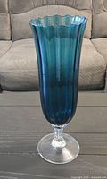 Full view of a tall teal glass vase with fluted sides and a clear glass pedestal base sitting on a table in front of a beige sofa.