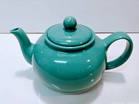 Turquoise ceramic teapot with a rounded body, curved spout, handle, and round lid with knob on top on a white background.