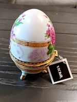 Porcelain egg-shaped trinket box with pink flowers and gold band, shown from an angled top view on a wooden surface.