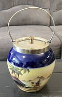 Full view of the biscuit barrel showing painted ceramic body with landscape and metal lid with handle.