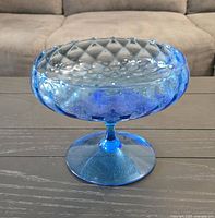 Single photo showing a blue glass decorative bowl with a textured diamond pattern and pedestal base on a wooden surface.