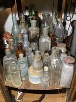 Image showing multiple antique glass bottles of various colors, shapes, and sizes arranged on wooden shelf under table. Bottles include clear, green, and amber glass with some having cork stoppers. Wear and age visible on glass surfaces.