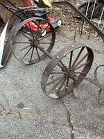 Photo showing entire pair of antique large metal wheels attached by a central axle on ground outdoors, rusted old condition.