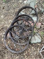Four antique, heavily rusted steel spoked wheels stacked on ground outdoors among leaves and stones.
