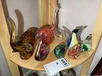 Photo showing a collection of seven colorful art glass animal figures displayed on a wooden curved shelf including elephant, swan, bird, and fish designs.