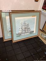 Two vintage framed ship prints by Ben Babelowsky leaning against wall on black surface. Both prints show detailed multi-masted sailing ships with light green matting and light wood frames with some wear.