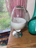 Front side view of the Brookstone candy dispenser showing clear egg-shaped container, white base and chrome stand, with a background window and pink curtain.