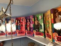 Seven Club 7 dolls displayed in their original rectangular packaging lined up on a ledge. Dolls include both males and females with colorful outfits and hairstyles, standing approximately 11 inches tall. Boxes have the distinctive red and white 'S CLUB 7' branding and rainbow stripe design on the edges.