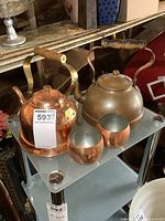 Photo showing two copper kettles on shelf, wooden handles, tarnished surfaces, with two copper cups in front