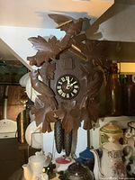 Front view of vintage wooden cuckoo clock showing black clock face with white Roman numerals, carved bird on top with damages on wing, carved leaf motifs surrounding clock face, weights and pendulum hanging below.
