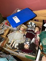 Box of assorted porcelain dolls in various clothing and styles including floral, plaid, and white dresses with detailed features and accessories