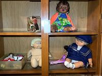 Various dolls including Cabbage Patch Kid, baby doll, Barbie-style doll and others displayed in wooden cabinet shelf