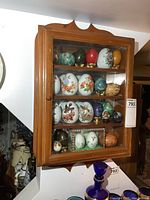 Front view of wooden display case with glass door, showing all decorative eggs inside arranged on three shelves.