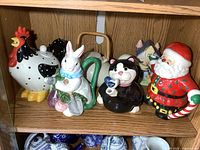 Front view of all six colorful, decorative ceramic teapots on a wooden shelf including rooster, rabbit, cat, duck, Santa, and cottage shapes