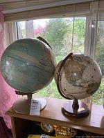 Two different globes on a table by a window, one with a white base and one with a wooden base