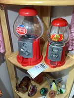 Two candy machines side by side on wooden shelf; left is red plastic Dubble Bubble with large clear plastic globe, right is red metal Jelly Belly machine with smaller glass globe