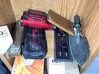 Multiple items including two vintage harmonicas, a harmonica case, two pens and small metal shovel door knocker placed on a wooden shelf.