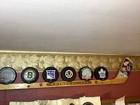 View of 7 NHL hockey pucks representing Original Six teams mounted or displayed on an 'Original Six' hockey stick shelf horizontally mounted on a wall.