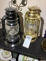 Full view of two metal lanterns, black and gold colored, standing side by side on shelf.