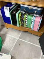Six lunch pails arranged on a shelf, showing a variety of colors and designs including a blue metal box, a green and black striped box, and a plastic Ninja Turtles pail.