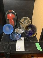 Six mixed glass paperweights displayed on dark shelf, showing variety of shapes, colors, and internal bubble patterns.