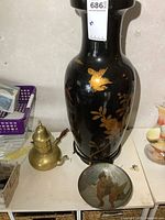 Photo of large black porcelain vase with gold bird motif along with brass teapot and brass bowl on white surface.