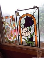 Two stained glass panels displayed hanging in a window, one with purple iris and white flower and the other with multi-color floral and leaf design.