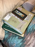 Stack of the framed stamps, stamp cover binder, and stamp collectors book on a bed.