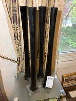Four tall black art glass vases standing on a glass surface near a window and curtain.