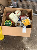 Box containing a variety of vintage tobacco tins with visible brand names including Amphora, Daily Mail, and others. The tins show signs of wear and aging.