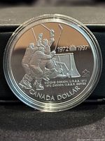 Obverse side of the silver proof coin with hockey players celebrating a goal near the net, dated 1972-1997 with inscriptions.