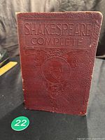 Front cover of the book embossed with 'SHAKESPEARE COMPLETE' and a medallion of Shakespeare's portrait with theatrical masks decoration.