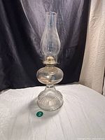 Full view of the vintage glass kerosene lamp showing ribbed embossed glass base, round oil globe, Eagle metal burner, and tall clear glass chimney against a black curtain background.