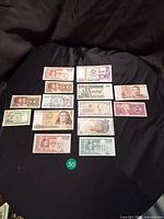 14 assorted uncirculated world paper money notes arranged on black fabric background. Notes feature various designs and denominations, including images of portraits and cultural symbols.