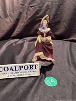 Front full view of the Coalport porcelain figurine of a Victorian lady wearing a deep red dress with yellow sleeves and blue bow at the neck against a black background.